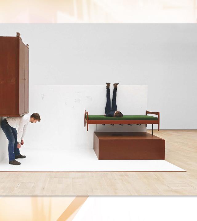 Das in einem großen Museumsraum ausgestellte Kunstwerk besteht aus einem Schrank, der verkehrt herum an der Wand hängt. Unter ihm beugt sich ein Mann vor, als ob er den Schrank auf dem Rücken tragen würde. An der Wand daneben hängt ein Bett, auf dem eine Person quer liegt und die Beine an der Wand hoch streckt. Darunter liebt ein Schrank quer.