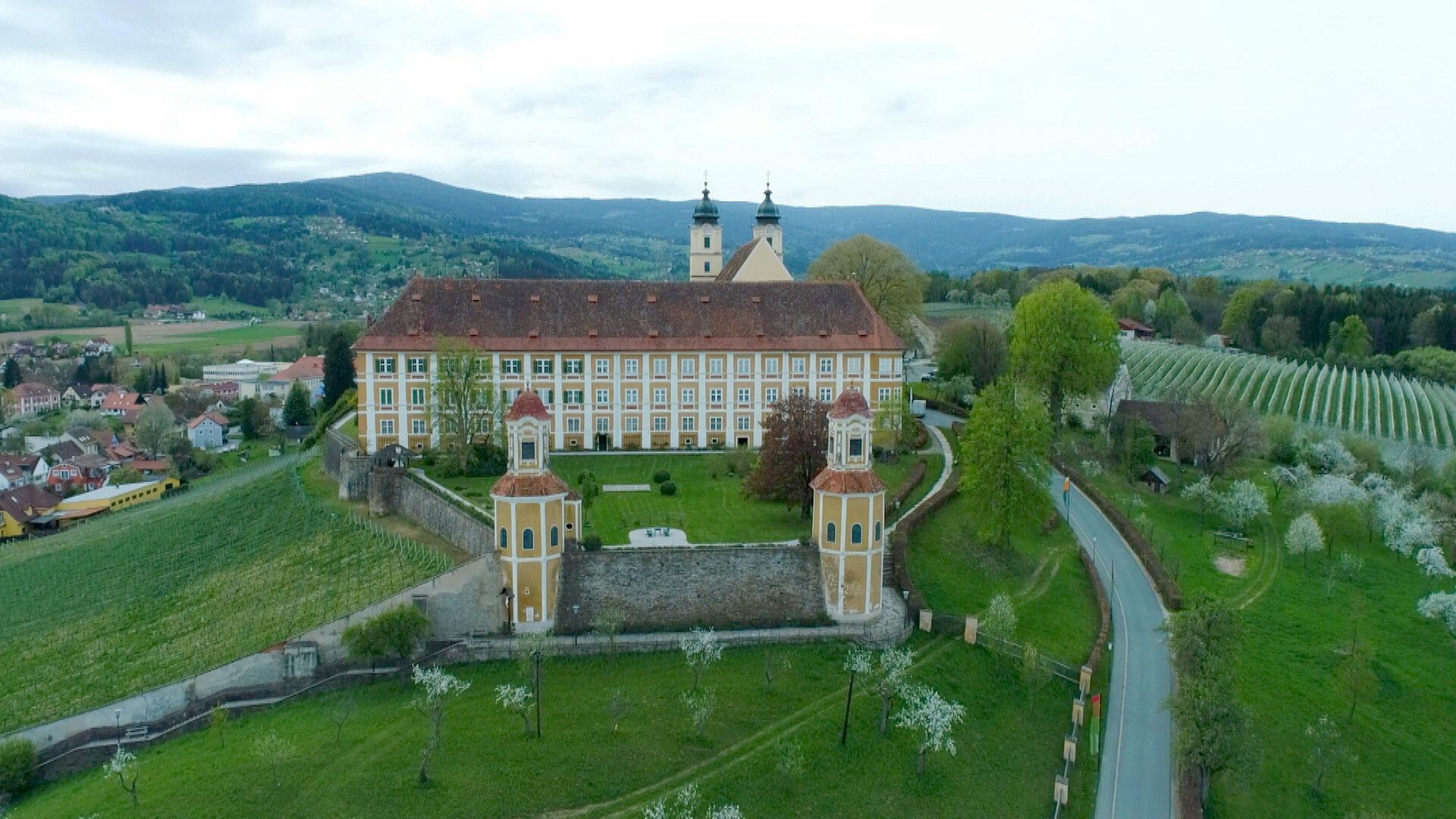Das Bild zeigt das Schloss Stainz, ein historisches Gebäude in der Steiermark, Österreich. Es hebt sich durch seine gelbe Fassade mit weißen Fensterrahmungen und ein rotes Ziegeldach hervor. Vor dem Schloss befinden sich zwei identische Türme mit runden Dächern, die farblich zur Fassade passen. Der Garten vor dem Schloss ist gepflegt und enthält einige Bäume und Sträucher.  Im Hintergrund erstrecken sich sanfte Hügel und Landschaften mit Weinreben, die in Reihen angeordnet sind. Die Umgebung wirkt grün und ländlich, passend zu einer ruhigen ländlichen Idylle. Ein Weg verläuft durch die Szene und führt in Richtung des Schlosses sowie in die umgebenden Felder. Der Himmel ist bewölkt, was auf wechselhaftes Wetter hindeutet.   Insgesamt vermittelt das Bild einen Eindruck von historischer Architektur in einer naturschönen Umgebung.