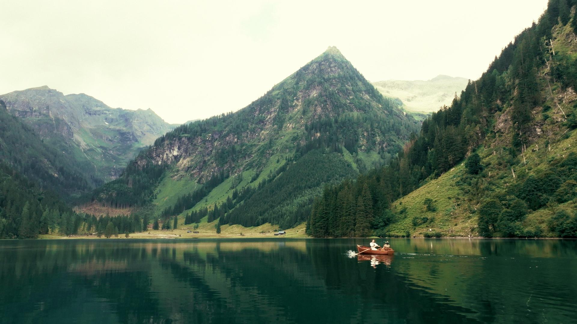 Das Bild zeigt eine idyllische Landschaft mit einem ruhigen, klaren See in der Mitte. Die Umgebung ist von steilen, bewaldeten Bergen umgeben, deren grüne Hänge bis zum Wasser hinunterführen. Die Berge sind teils mit Felsen und teils mit dichten Wäldern bedeckt, die sich über die Hänge verteilen. Am Ufer des Sees sind einige Gebäude, vermutlich Almhütten, erkennbar.  Auf dem Wasser ist ein kleines, hölzernes Boot zu sehen, in dem zwei Personen sitzen. Sie rudern sanft über die spiegelglatte Oberfläche des Sees. Der Himmel ist bewölkt, was dem Bild eine sanfte, gedämpfte Lichtatmosphäre verleiht.  Insgesamt vermittelt die Szene ein Gefühl von Ruhe und Naturverbundenheit, unterstrichen durch die majestätischen Berge und die stille Wasseroberfläche.