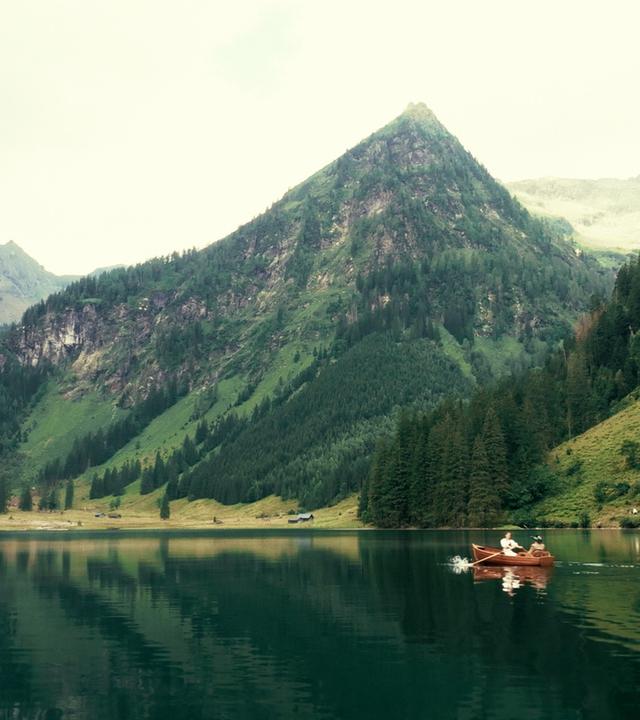 Das Bild zeigt eine idyllische Landschaft mit einem ruhigen, klaren See in der Mitte. Die Umgebung ist von steilen, bewaldeten Bergen umgeben, deren grüne Hänge bis zum Wasser hinunterführen. Die Berge sind teils mit Felsen und teils mit dichten Wäldern bedeckt, die sich über die Hänge verteilen. Am Ufer des Sees sind einige Gebäude, vermutlich Almhütten, erkennbar.  Auf dem Wasser ist ein kleines, hölzernes Boot zu sehen, in dem zwei Personen sitzen. Sie rudern sanft über die spiegelglatte Oberfläche des Sees. Der Himmel ist bewölkt, was dem Bild eine sanfte, gedämpfte Lichtatmosphäre verleiht.  Insgesamt vermittelt die Szene ein Gefühl von Ruhe und Naturverbundenheit, unterstrichen durch die majestätischen Berge und die stille Wasseroberfläche.