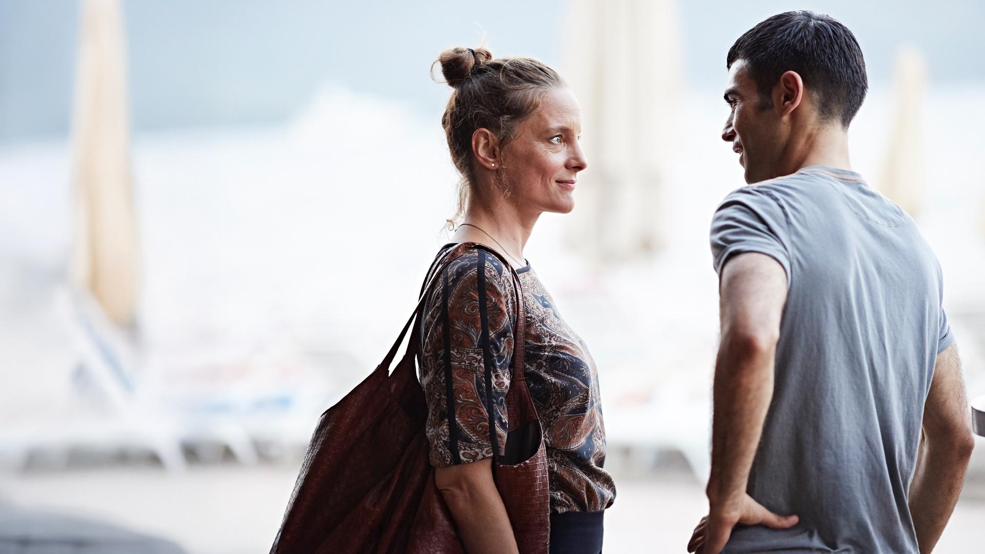 Marion (Anne Ratte-Polle) und Baran (Ogulcan Arman Uslu) stehen sich gegenüber und lächeln sich an. Im Hintergrund ist ein türkischer Urlaubsort zu erahnen.