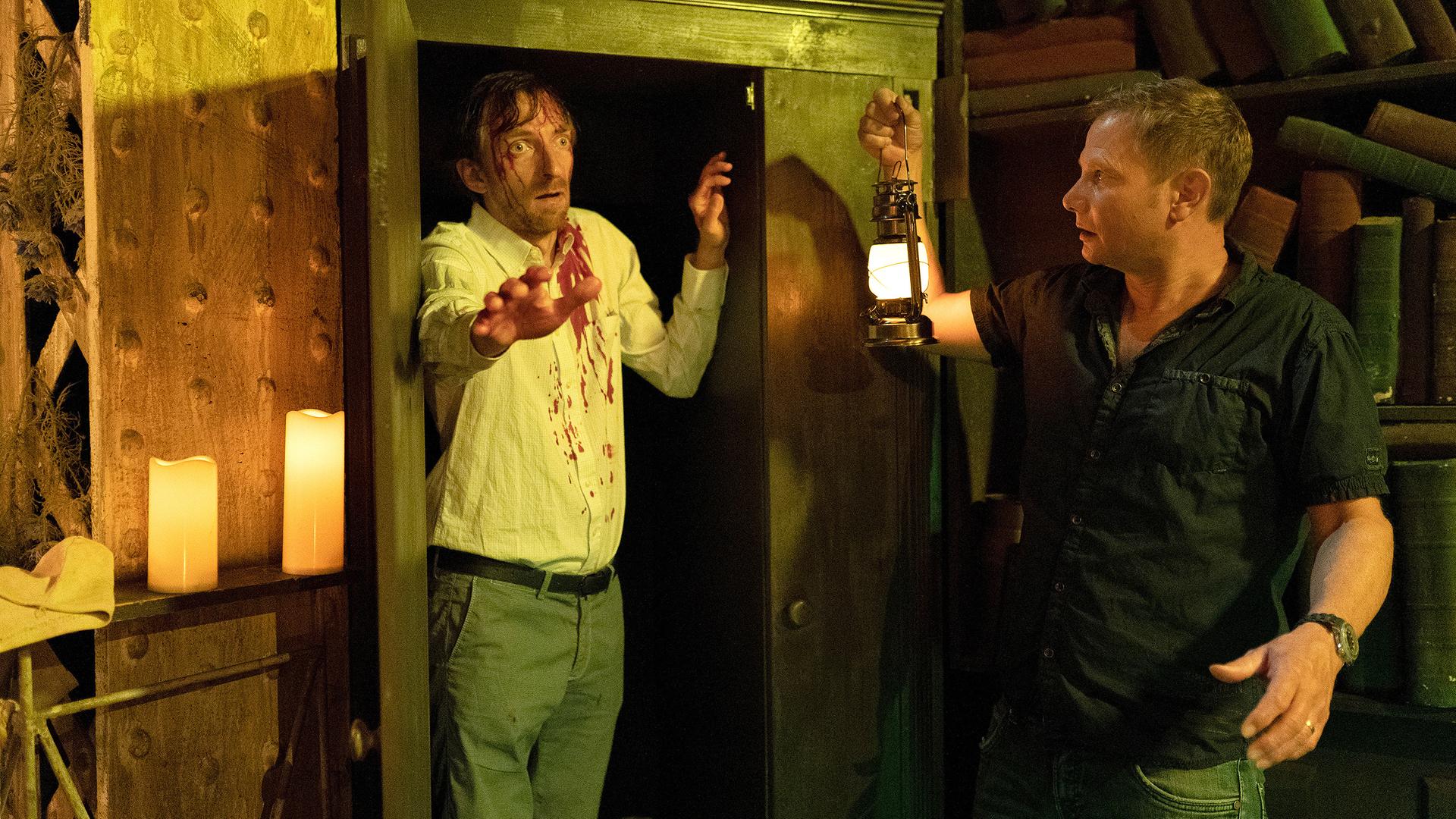 Haase (Fabian Harloff) steht mit einer Gaslampe in der rechten Hand vor einem Wandschrank. In der linken Tür des Schranks steht ein Mann (Hendrik von Bültzingslöwen) mit weit aufgerissenen Augen. Auf seinem weißen Hemd sind Blutflecken zu sehen. Seine rechte Hand hat er nach vorne ausgestreckt. Haase schaut erschrocken.