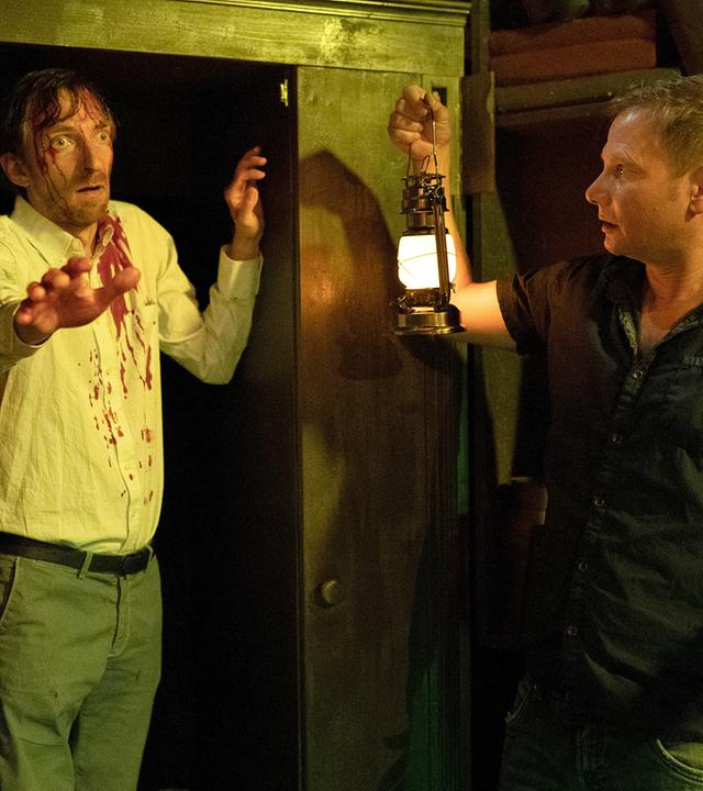 Haase (Fabian Harloff) steht mit einer Gaslampe in der rechten Hand vor einem Wandschrank. In der linken Tür des Schranks steht ein Mann (Hendrik von Bültzingslöwen) mit weit aufgerissenen Augen. Auf seinem weißen Hemd sind Blutflecken zu sehen. Seine rechte Hand hat er nach vorne ausgestreckt. Haase schaut erschrocken.