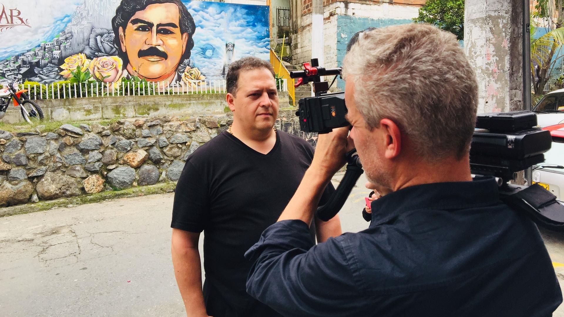 Juan Pablo Escobar steht vor einer Mauer. Auf der Mauer befindet sich ein gemaltes Porträt von Pablo Escobar. Ein Mann richtet eine Videokamera auf Juan Pablo, der mit ernster Miene direkt in die Kamera blickt.