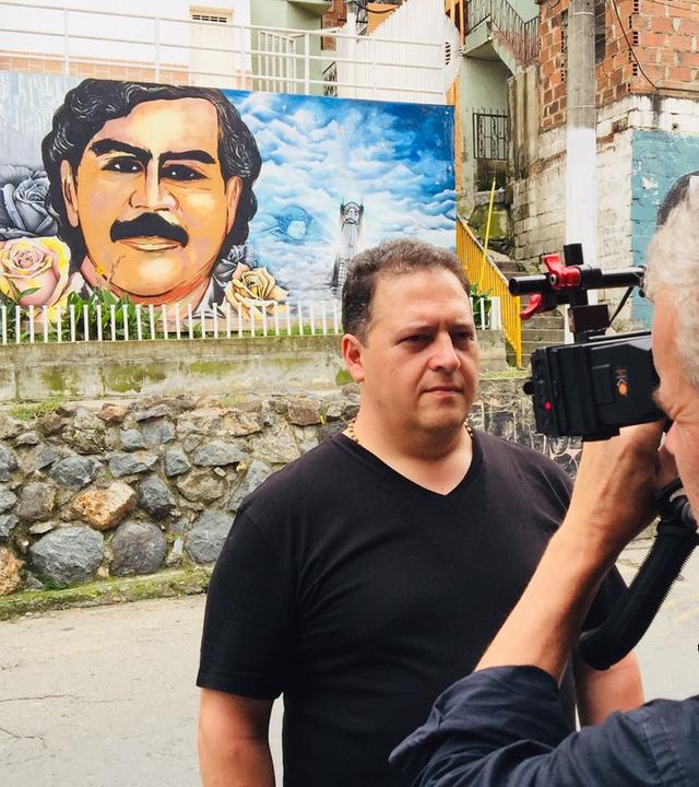 Juan Pablo Escobar steht vor einer Mauer. Auf der Mauer befindet sich ein gemaltes Porträt von Pablo Escobar. Ein Mann richtet eine Videokamera auf Juan Pablo, der mit ernster Miene direkt in die Kamera blickt.