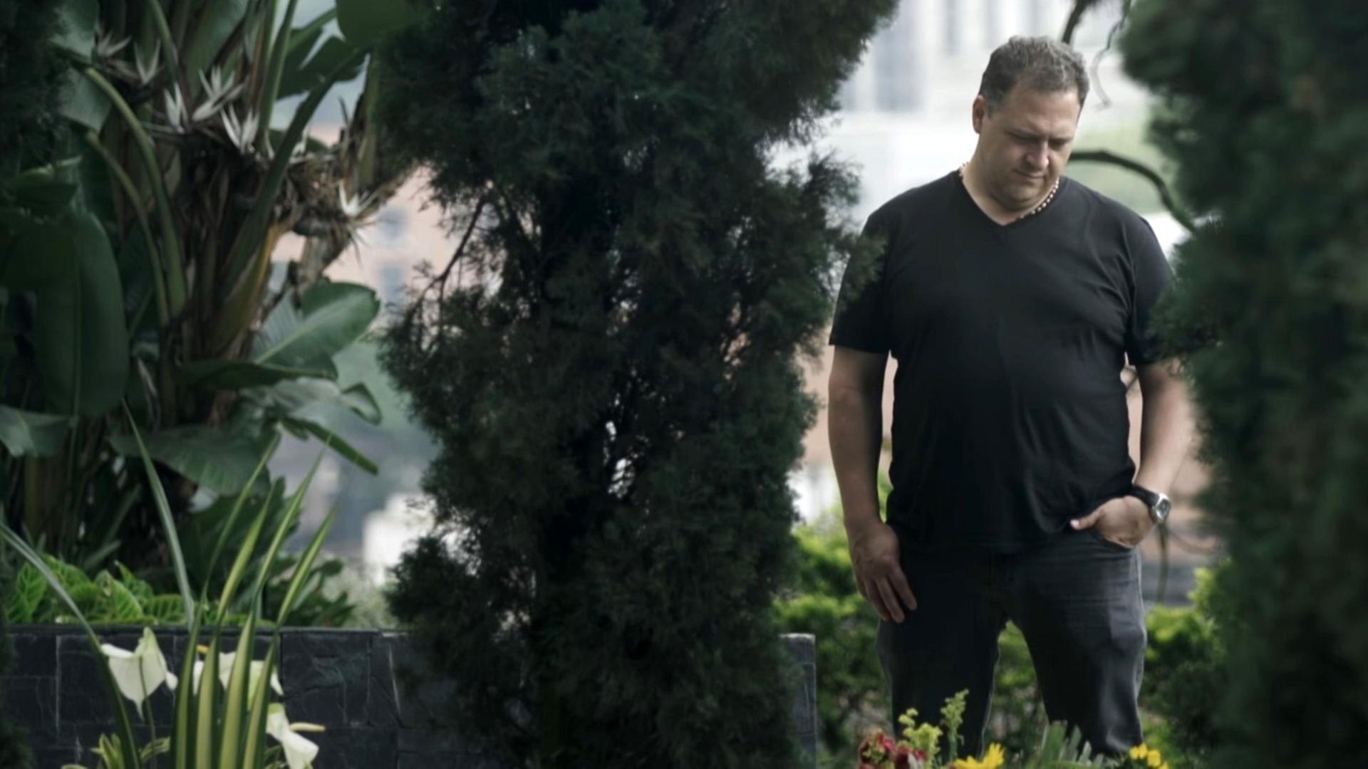 Juan Pablo Escobar steht auf einem Friedhof. Er blickt auf das Grab seines Vaters. Er ist schwarz gekleidet.