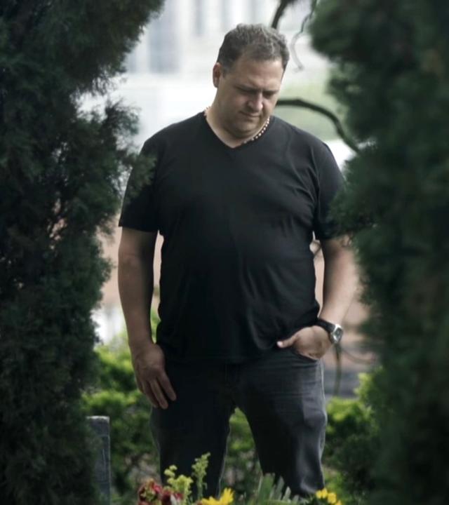 Juan Pablo Escobar steht auf einem Friedhof. Er blickt auf das Grab seines Vaters. Er ist schwarz gekleidet.