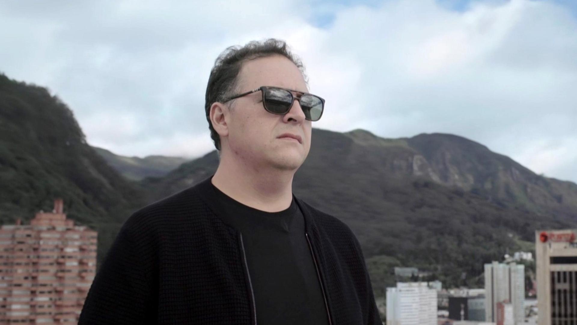 Aufnahme von Juan Pablo Escobar. Er hat kurze, ergrauende Haare, trägt eine dunkle Sonnenbrille und schwarze Kleidung. Hinter ihm sind Hochhäuser und Hügel zu sehen.