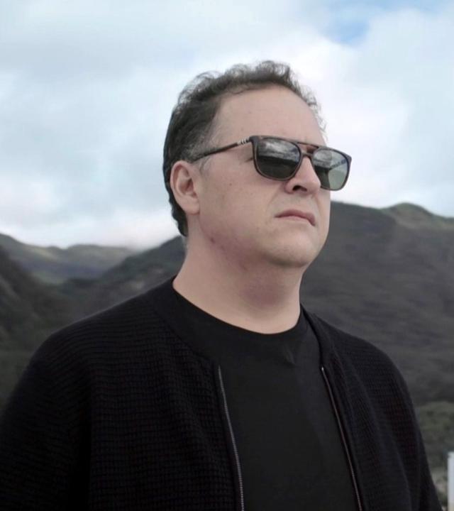 Aufnahme von Juan Pablo Escobar. Er hat kurze, ergrauende Haare, trägt eine dunkle Sonnenbrille und schwarze Kleidung. Hinter ihm sind Hochhäuser und Hügel zu sehen.