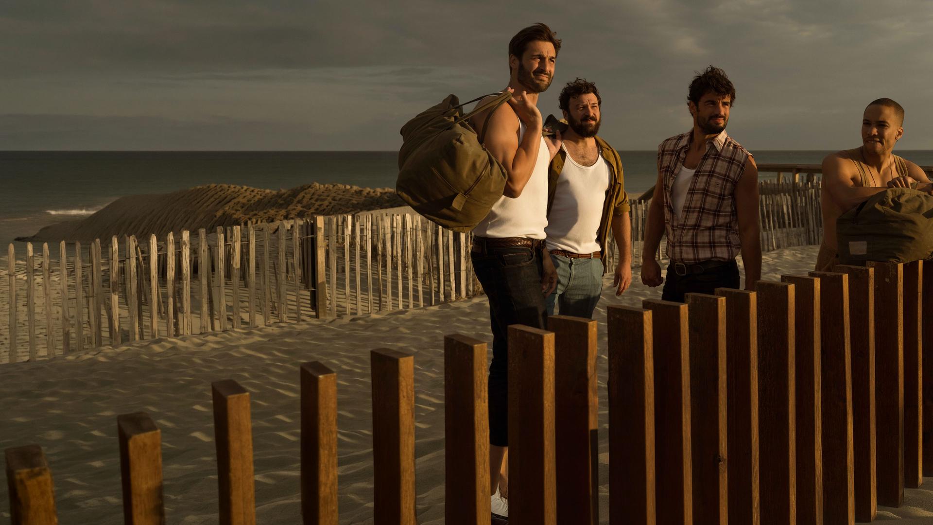 "Escort Boys - Imkerei mit Zusatzleistungen": Ben (Guillaume Labbé), Ludo (Thibaut Evrard), Mathias (Simon Ehrlacher) und Zak (Corentin Fila) stehen vor einem braunen Holzzaun am Meer. Ben und Zak haben große Taschen bei sich: Ben trägt seine Tasche über der linken Schulter, Zak hat seine auf dem Zaun abgestellt und stützt sich mit verschränkten Armen darauf. Alle vier tragen leichte Kleidung und blicken konzentriert. Sonnenlicht blendet ihre Gesichter leicht.