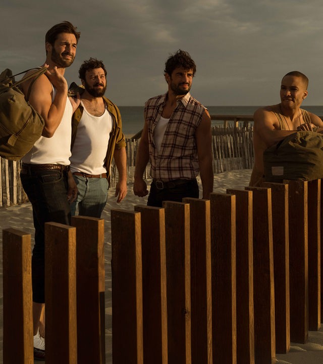 "Escort Boys - Imkerei mit Zusatzleistungen": Ben (Guillaume Labbé), Ludo (Thibaut Evrard), Mathias (Simon Ehrlacher) und Zak (Corentin Fila) stehen vor einem braunen Holzzaun am Meer. Ben und Zak haben große Taschen bei sich: Ben trägt seine Tasche über der linken Schulter, Zak hat seine auf dem Zaun abgestellt und stützt sich mit verschränkten Armen darauf. Alle vier tragen leichte Kleidung und blicken konzentriert. Sonnenlicht blendet ihre Gesichter leicht.