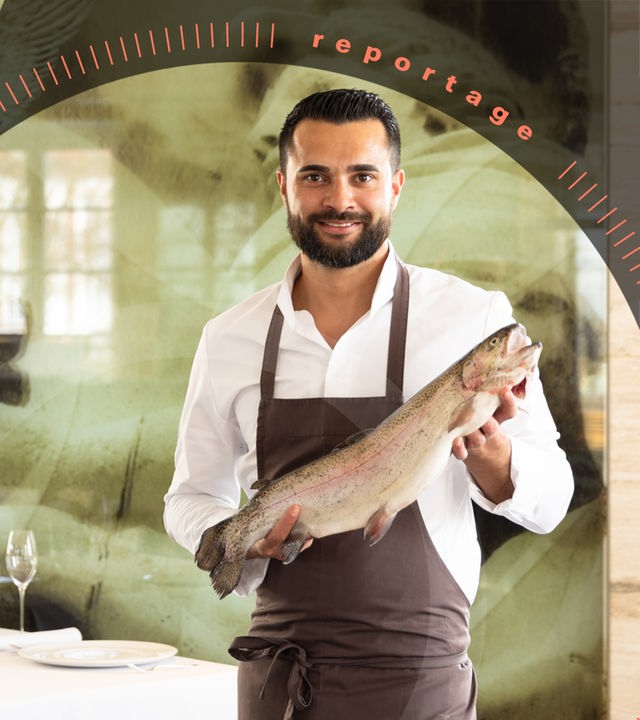 Küchenchef Dennis Kuckuck hält einen großen Fisch in der Hand und lächelt in die Kamera. Er trägt eine Schürze. Im Hintergrund ist der schick gedeckte Tisch eines Restaurants zu sehen.