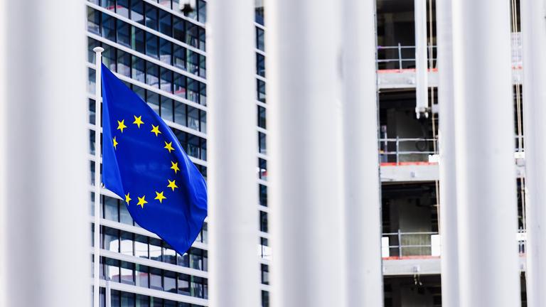 Eine Flagge der Europäischen Union (EU) weht vor dem Gebäude des Europäischen Parlaments und ist zwischen anderen Fahnenmasten hindurch zu sehen. 
