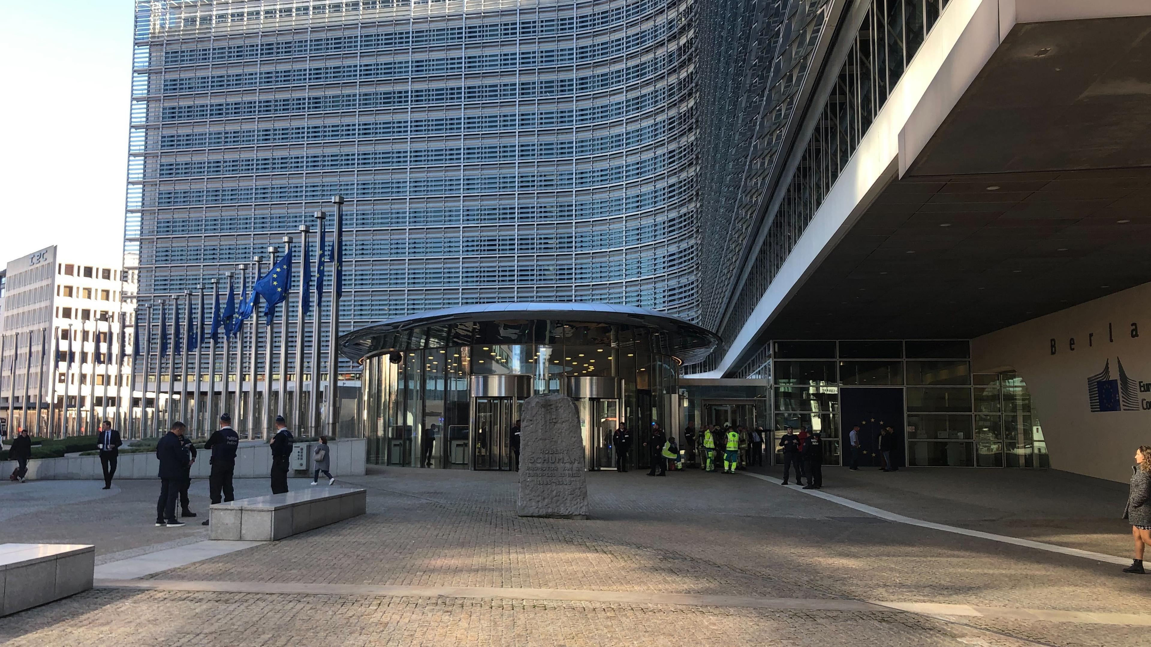 Die Sonne wirft Schatten vor dem modernen Gebäude der EU-Kommission. Wenige Menschen stehen auf dem Platz davor, neben EU-Flaggen.