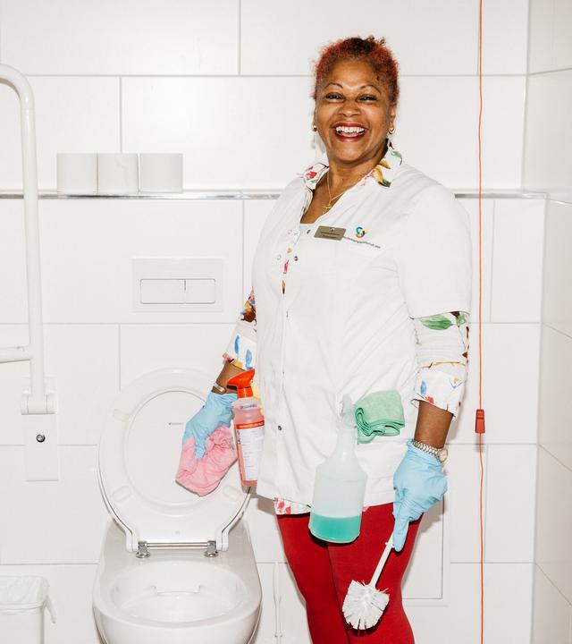 Jenny steht vor einer Toilette und schaut lachend in die Kamera. In einer Hand hält sie ein Putztuch in der anderen eine Toilettenbürste.