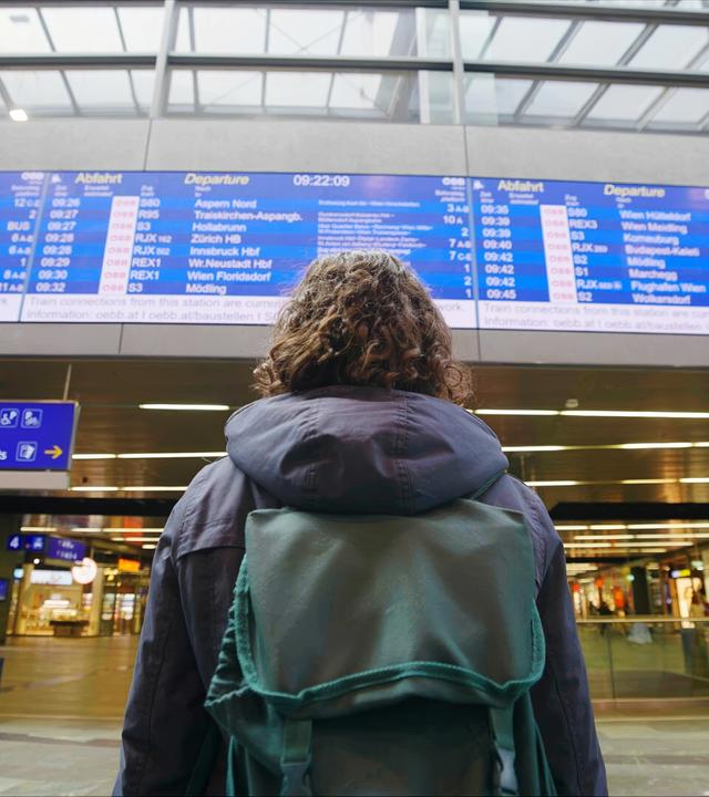 Das Bild zeigt eine Person mit lockigem Haar, die in einer modernen, hellen Bahnhofshalle steht. Die Person trägt eine grüne Rucksacktasche und schaut auf eine große Anzeigetafel über ihr. Diese Tafel zeigt die Abfahrtszeiten von Zügen mit Zielen, die teilweise in deutscher Sprache vermerkt sind. Die Halle hat große Fenster, durch die Tageslicht strömt, und eine offene, zugängliche Architektur. Auf dem Boden sind andere Reisende zu sehen, die mit Gepäck in verschiedenen Richtungen gehen. An den Wänden sind Informationsschilder, und es gibt einen Bereich mit Ticketautomaten. Insgesamt vermittelt die Umgebung ein geschäftiges Reisewohlgefühl.