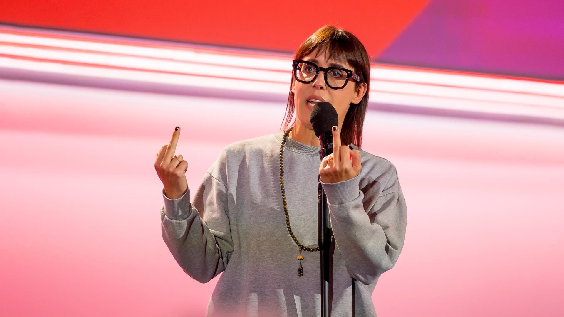Eva Karl Faltermeier trägt einen grauen Pullover. Sie zeigt beide Mittelfinger. Sie trägt eine Brille. Der Hintergrund ist hell und rot beleuchtet.