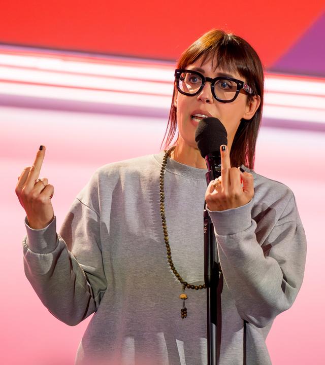 Eva Karl Faltermeier trägt einen grauen Pullover. Sie zeigt beide Mittelfinger. Sie trägt eine Brille. Der Hintergrund ist hell und rot beleuchtet.
