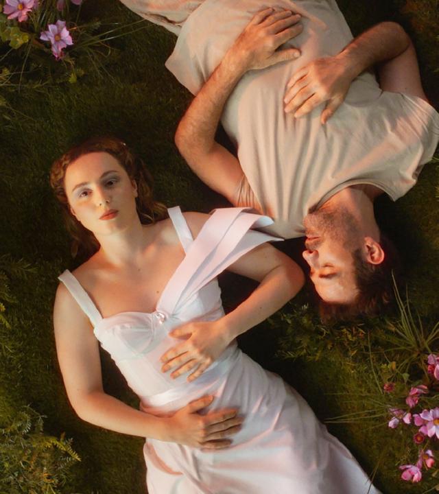 Das Bild zeigt eine Szene aus der Vogelperspektive, in der eine Frau und ein Mann nebeneinander auf einer grünen, moosbewachsenen Fläche liegen. Die Umgebung ist mit zarten, pinken Blumen übersät. Die Frau trägt ein elegantes, weißes Kleid mit breiten Trägern und blickt ruhig und ernst direkt in die Kamera. Der Mann neben ihr trägt ein einfaches, helles Oberteil und hat die Augen geschlossen. 