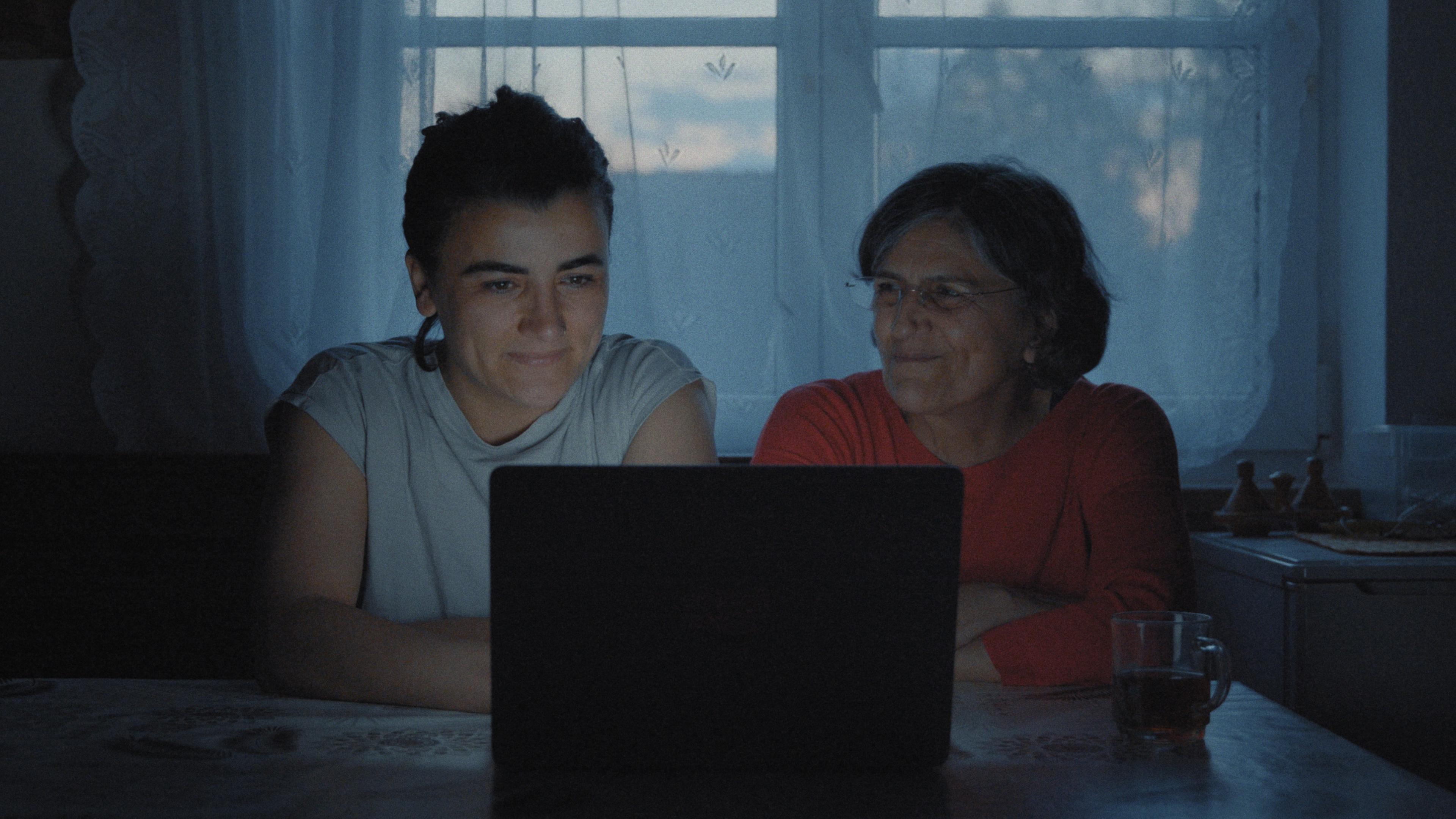 "Exile Never Ends": Mutter Yildiz Bektaş und Tochter Bahar Bektaş schauen lächelnd auf einen Laptop, der auf einem Küchentisch vor ihnen steht.