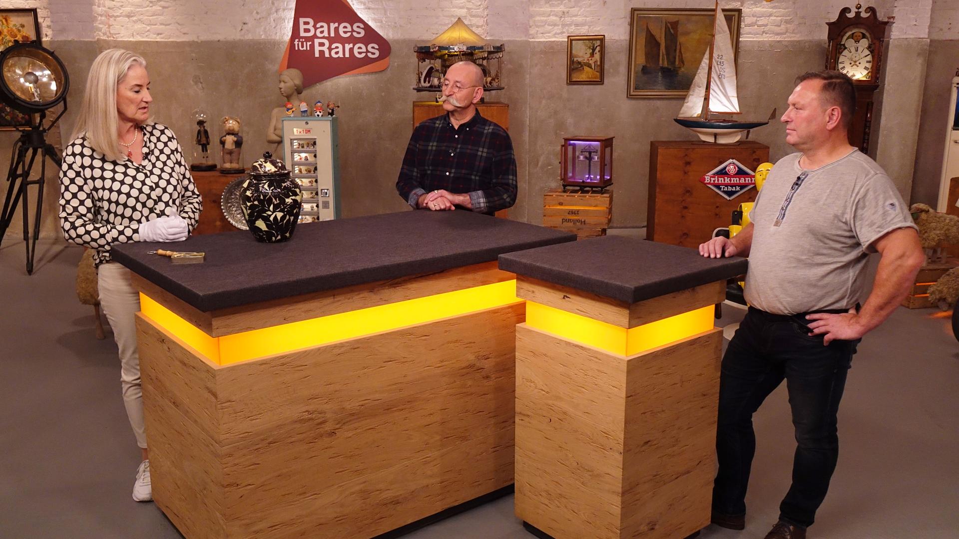 Expertin Dr. Friederike Werner (links) hat eine Deckelvase vor sich auf dem Tisch stehen und betrachtet sie. Horst Lichter (Mitte) und der Verkäufer (rechts) schauen sie an.