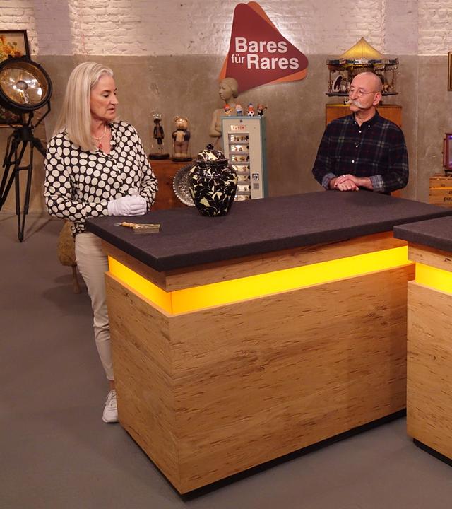 Expertin Dr. Friederike Werner (links) hat eine Deckelvase vor sich auf dem Tisch stehen und betrachtet sie. Horst Lichter (Mitte) und der Verkäufer (rechts) schauen sie an.