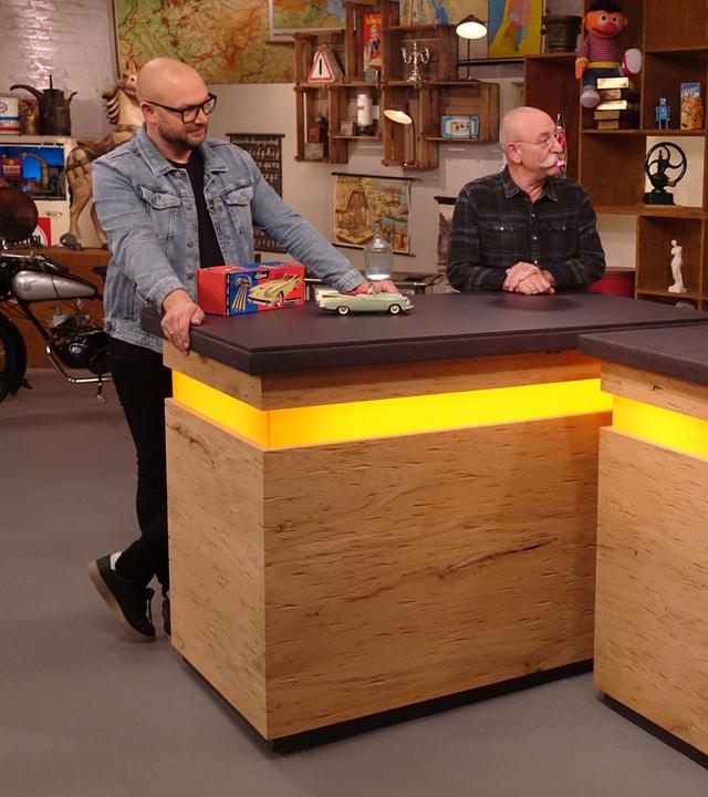 Experte Sven Deutschmanek (links) hat ein Fernlenkauto vor sich auf dem Tisch stehen. Er und Horst Lichter (Mitte) schauen das Verkäuferpaar (rechts) an.