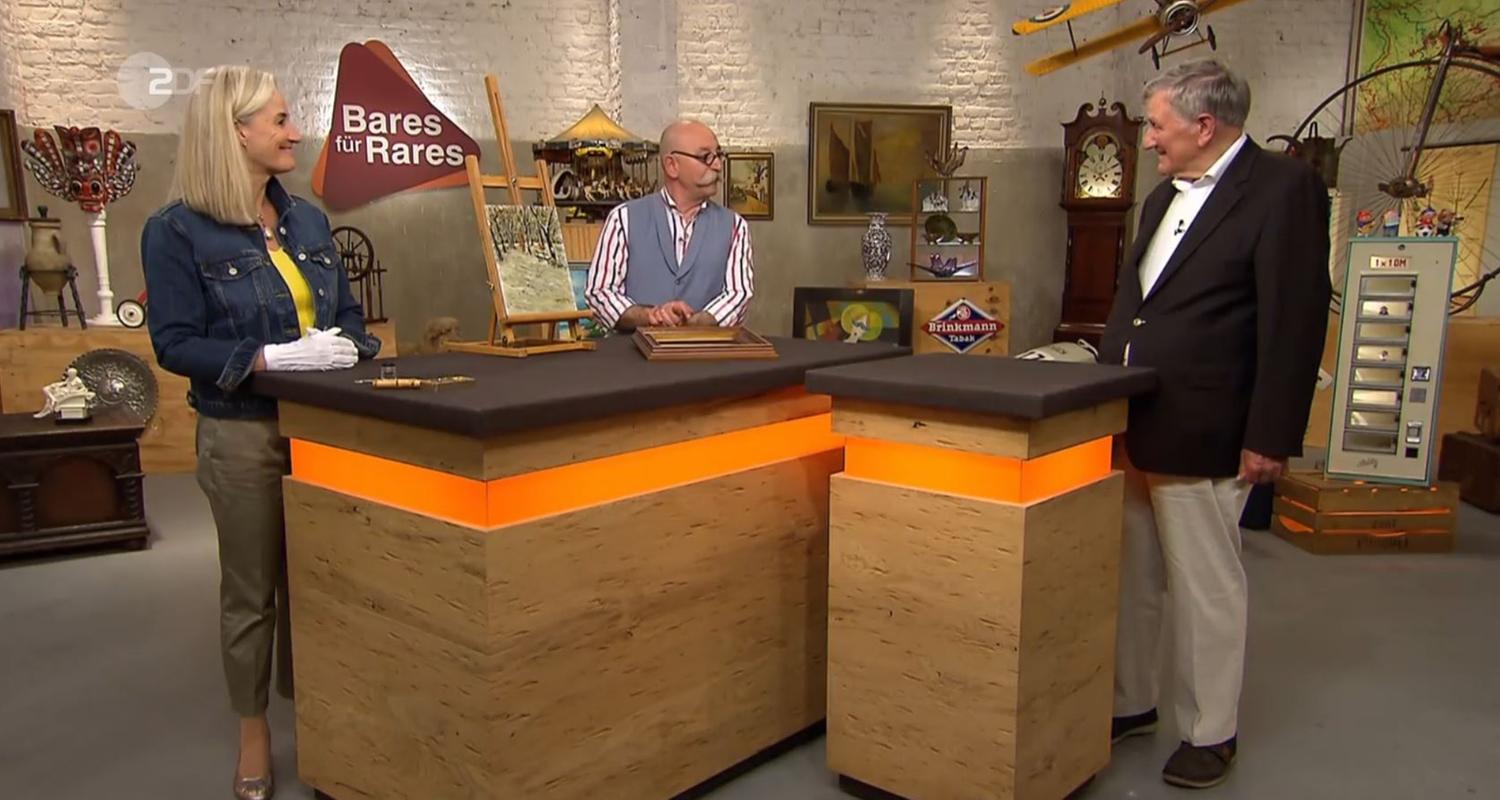 Expertin Dr. Friederike Werner steht links neben einem kleinen Gemälde, das auf einer kleinen Staffellei drapierten auf dem Expertisentisch steht. Sie und Horst Lichter (Mitte) schauen den Verkäufer (rechts) an.