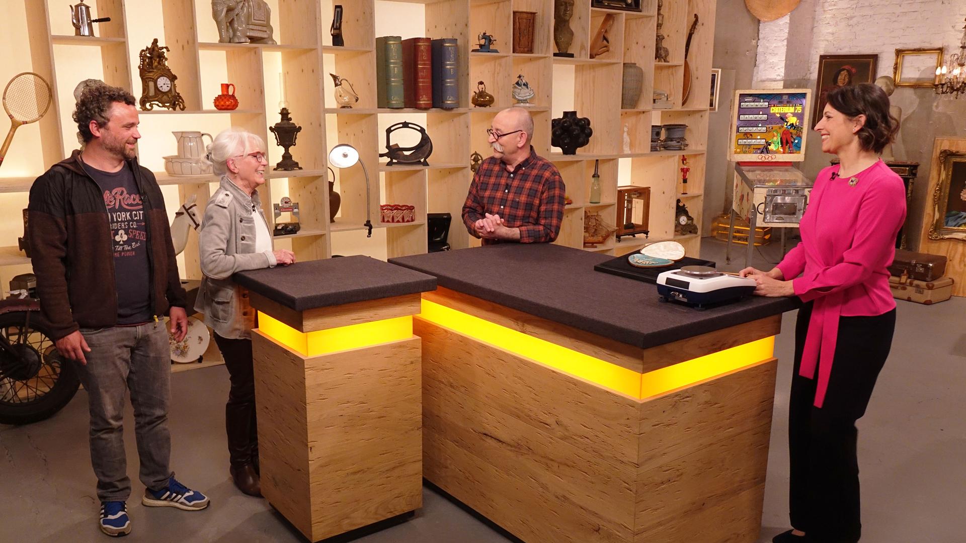 Wendela Horz steht in einer pinken Bluse am Expertentisch. Sie schaut zu einem Mann und einer Frau, die am Verkaufstisch stehen. In der Mitte steht Horst Lichter.