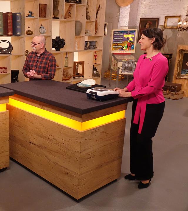 Wendela Horz steht in einer pinken Bluse am Expertentisch. Sie schaut zu einem Mann und einer Frau, die am Verkaufstisch stehen. In der Mitte steht Horst Lichter.