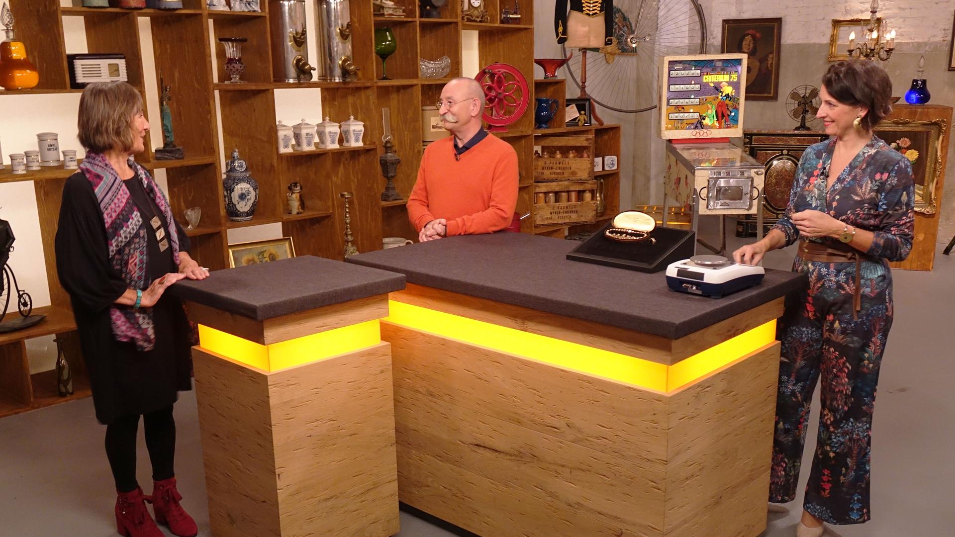 Expertin Wendela Horz (rechts) hat Schmuck und eine Waage vor sich auf dem Tisch stehen. Sie und Horst Lichter (Mitte) schauen die Verkäuferin (links) an.