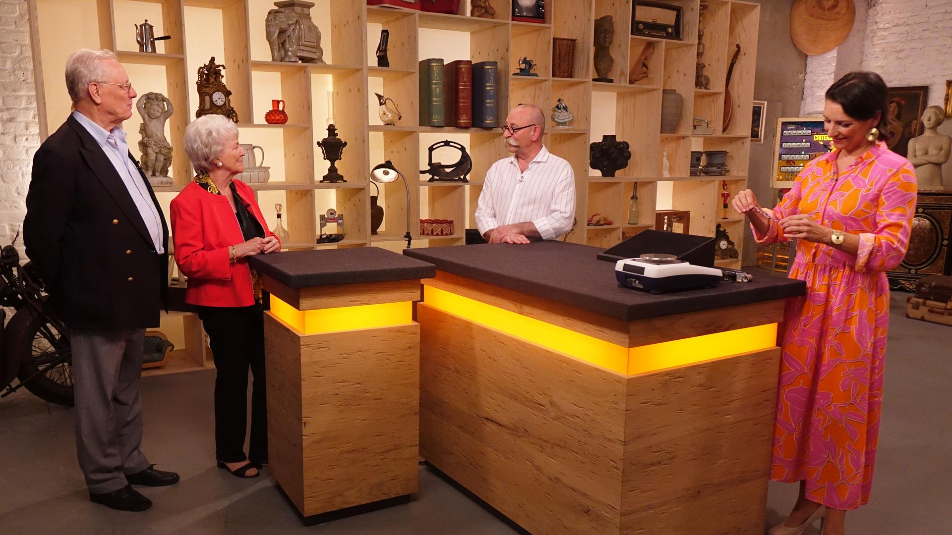 Expertin Wendela Horz (rechts) steht am Expertentisch, hält ein Schmuckstück in der Hand und betrachtet es. Horst Lichter (Mitte) spricht mit dem Verkäufer und der Verkäuferin (links).