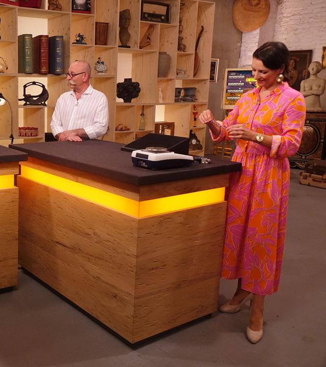 Expertin Wendela Horz (rechts) steht am Expertentisch, hält ein Schmuckstück in der Hand und betrachtet es. Horst Lichter (Mitte) spricht mit dem Verkäufer und der Verkäuferin (links).