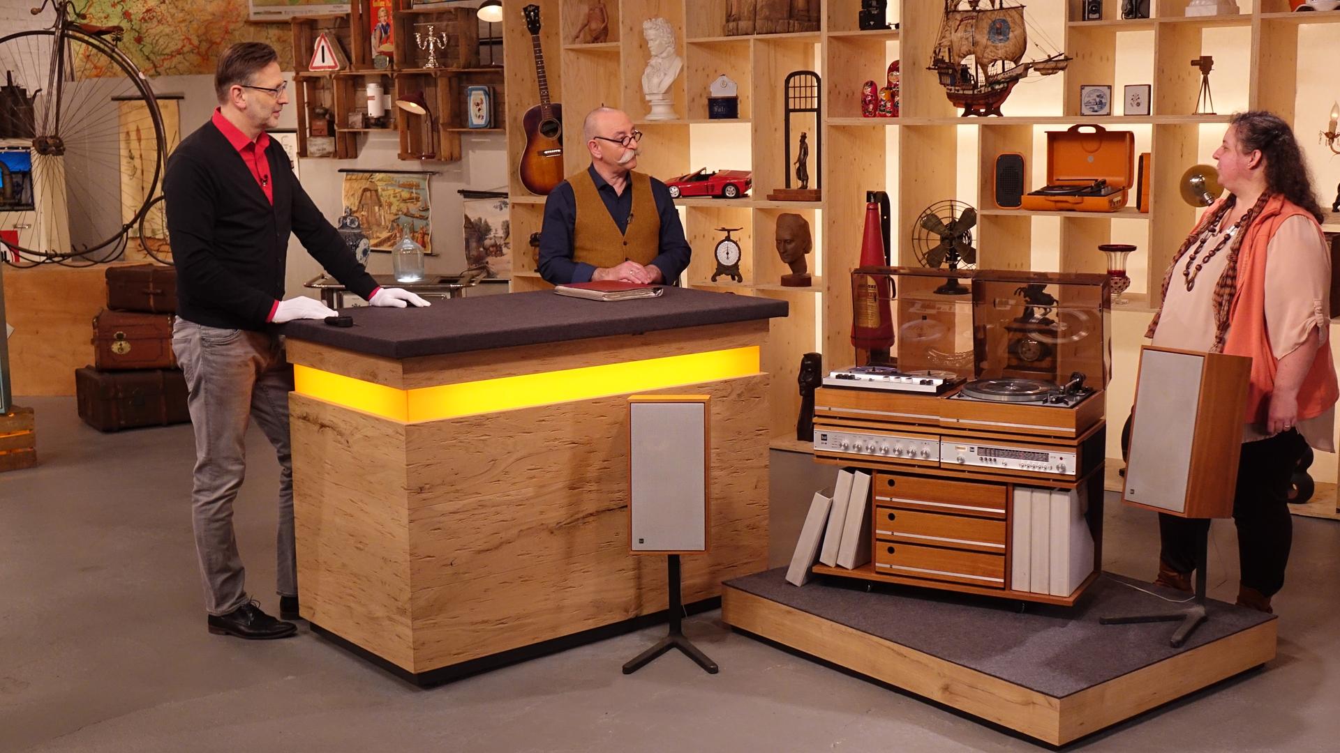 Experte Detlev Kümmel steht links neben Horst Lichter hinter dem Expertisentisch. Vor dem Tisch steht eine alte Stereoanlage mit Holzelementen und freistehenden Lautsprechern. Dahinter rechts steht die Verkäuferin.