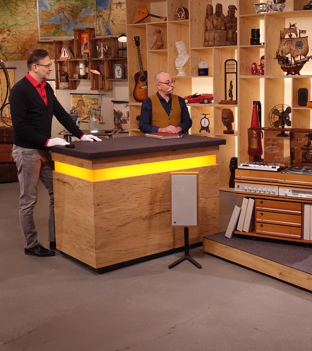 Experte Detlev Kümmel steht links neben Horst Lichter hinter dem Expertisentisch. Vor dem Tisch steht eine alte Stereoanlage mit Holzelementen und freistehenden Lautsprechern. Dahinter rechts steht die Verkäuferin.