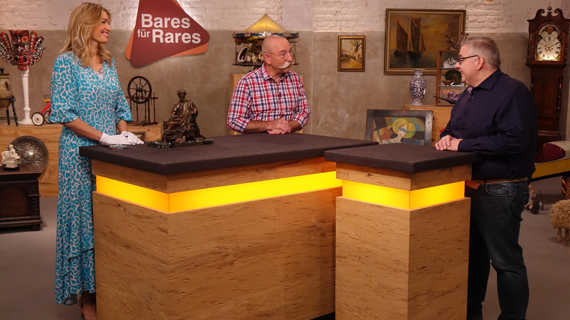 Expertise von Dr. Bianca Berding (links) zu einer Bronzeskulptur, die eine sitzende Person zeigt und neben ihr auf dem Tisch steht. Sie und Horst Lichter (Mitte) sehen den Verkäufer (rechts) an.