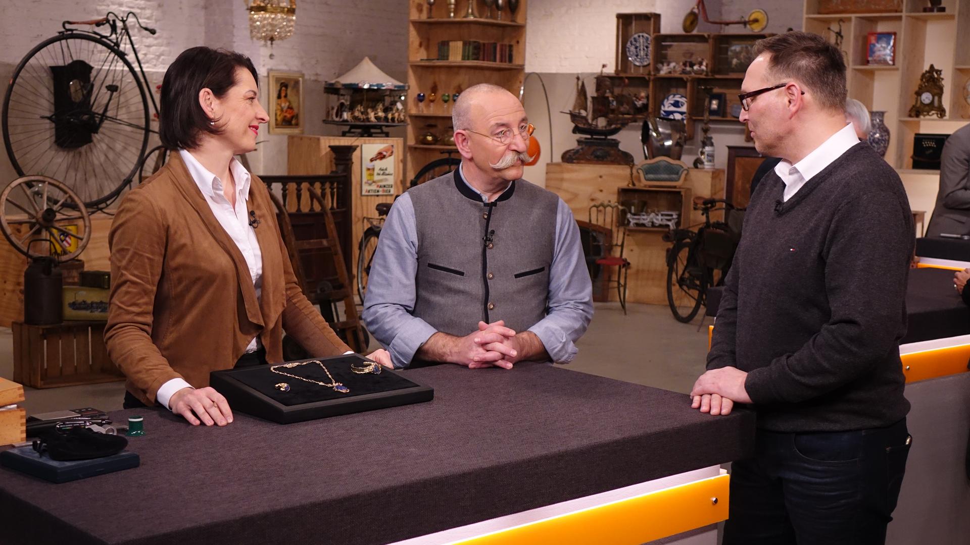 Expertin Wendela Horz (links) hat ein Schmuckset vor sich auf dem Tisch stehen. Sie und Horst Lichter (Mitte) schaut den Verkäufer (rechts) an.