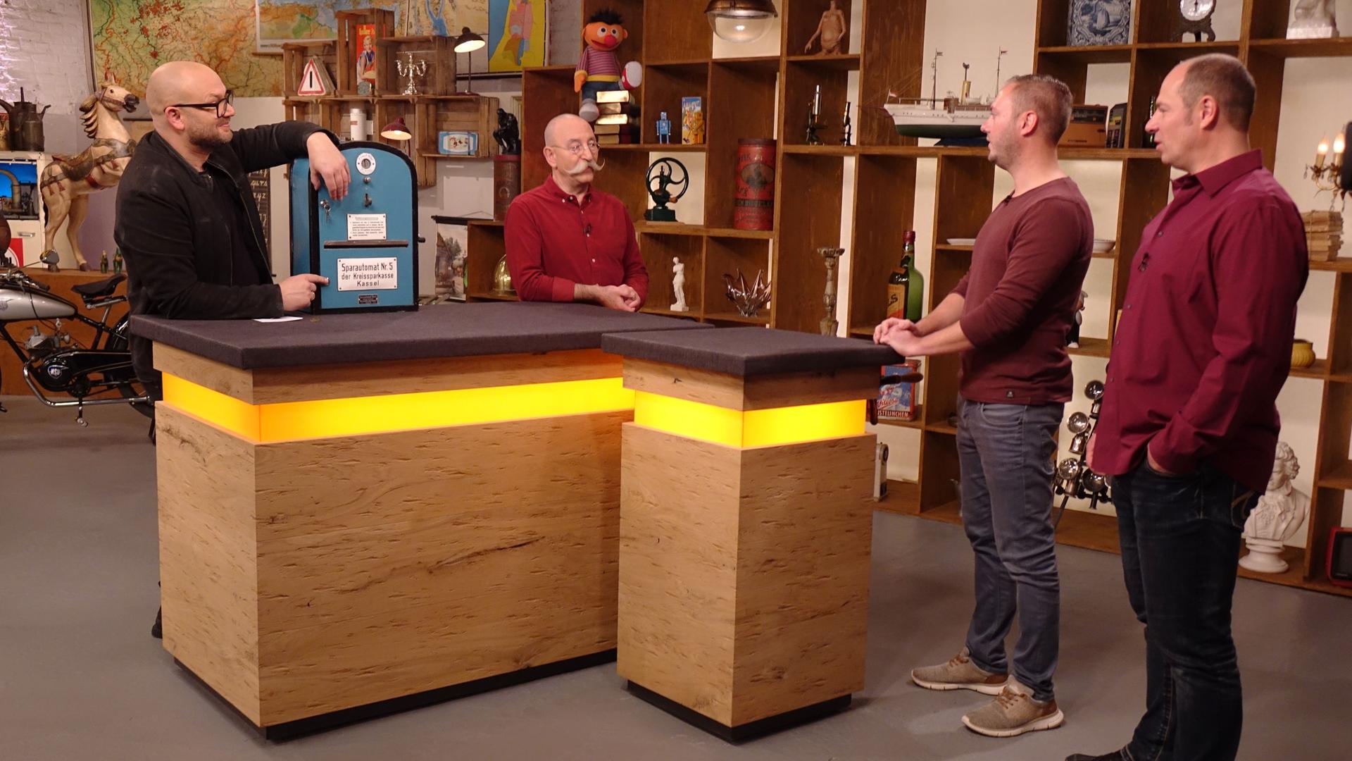 Experte Sven Deutschmanek (links) hat einen Automaten vor sich auf dem Tisch stehen. Er und Horst Lichter (Mitte) schauen die Verkäufer (rechts) an.