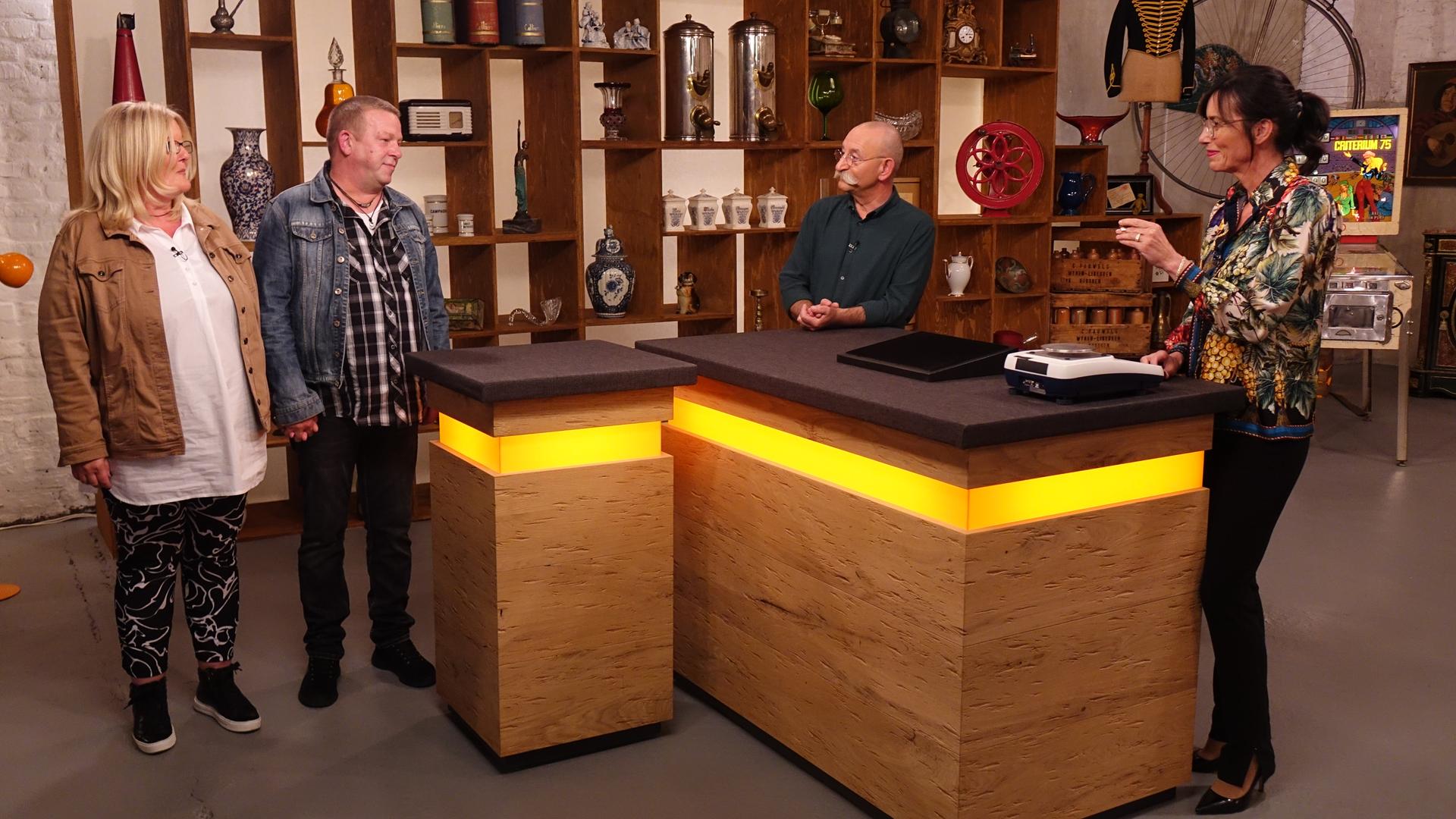 Expertin Dr. Heide Rezepa-Zabel (rechts) überprüft ein Schmuckstück an einer Waage. Sie und Horst Lichter (Mitte) schauen die beiden Verkäufer (links) an.