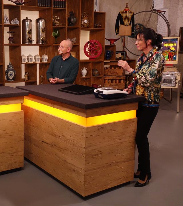 Expertin Dr. Heide Rezepa-Zabel (rechts) überprüft ein Schmuckstück an einer Waage. Sie und Horst Lichter (Mitte) schauen die beiden Verkäufer (links) an.