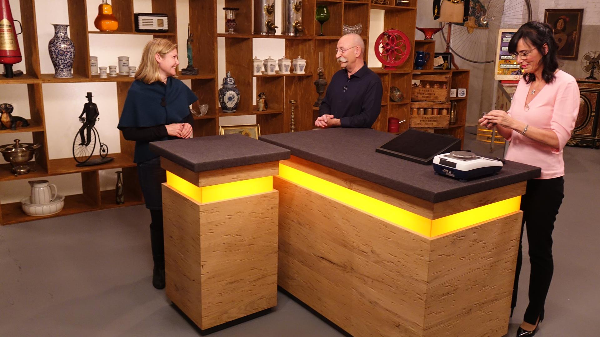 Expertin Dr. Heide Rezepa-Zabel (rechts) überprüft ein Schmuckstück an einer Waage, während Horst Lichter (Mitte) mit der Verkäuferin (links) spricht.