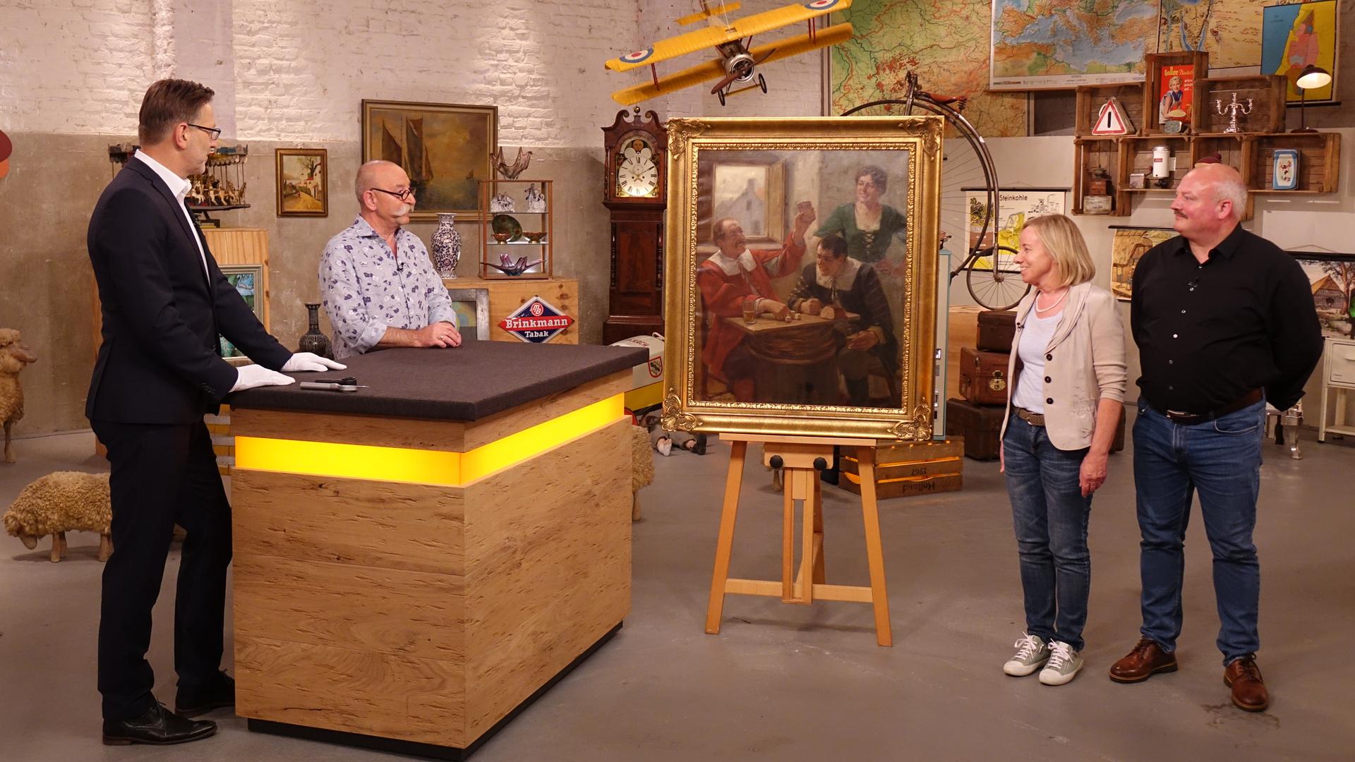 Experte Detlev Kümmel und Horst Lichter (links am Expertentisch) betrachten ein Gemälde in einem goldenen Rahmen, das auf einer Staffelei steht und drei Menschen in einem historischen Gasthaus zeigt. Rechts des Gemäldes stehen der Verkäufer und die Verkäuferin.