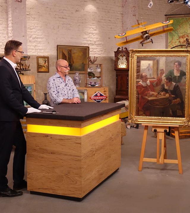Experte Detlev Kümmel und Horst Lichter (links am Expertentisch) betrachten ein Gemälde in einem goldenen Rahmen, das auf einer Staffelei steht und drei Menschen in einem historischen Gasthaus zeigt. Rechts des Gemäldes stehen der Verkäufer und die Verkäuferin.