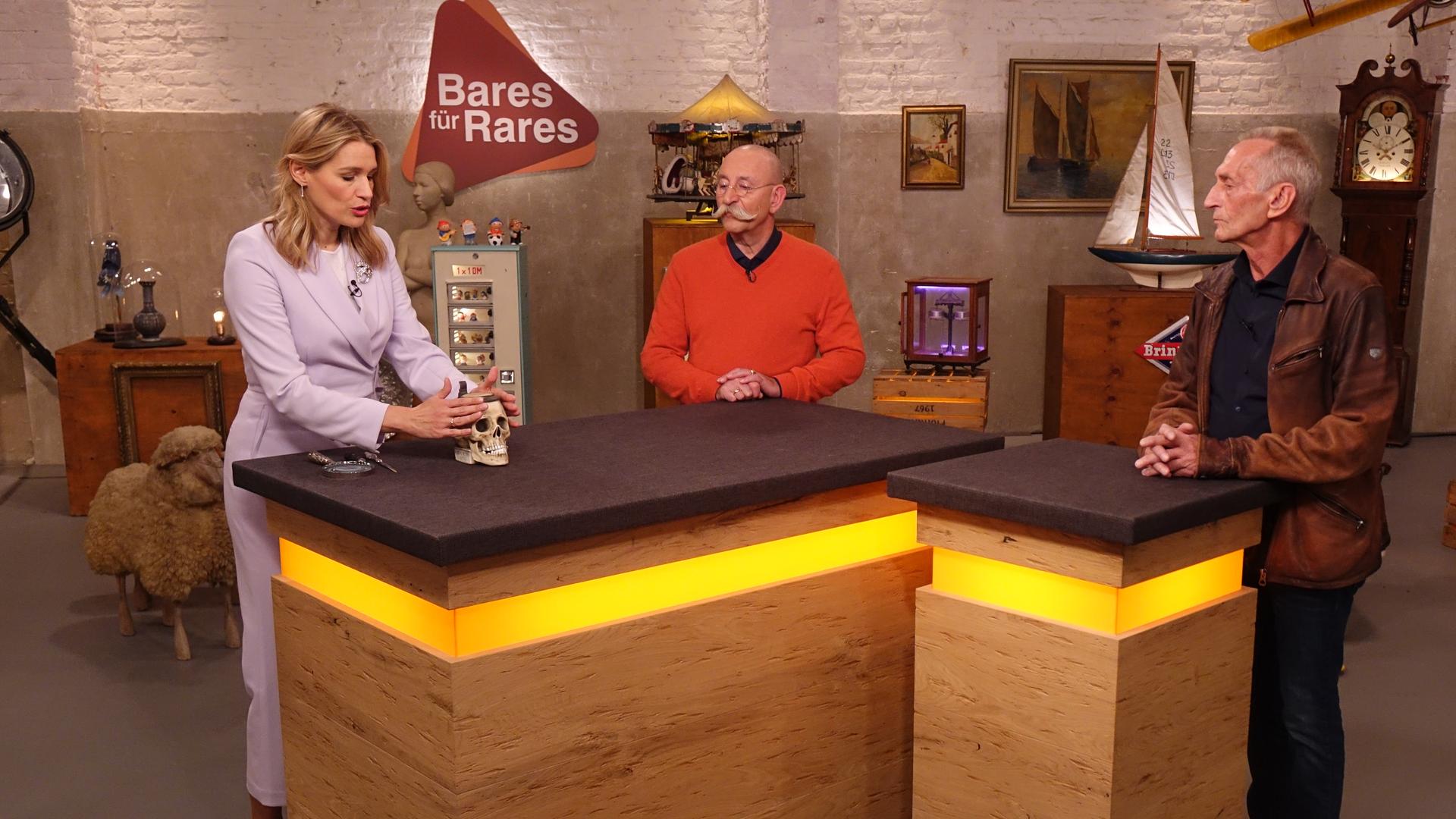 Expertin Dr. Bianca Berding (links) hat einen Bierkrug in Form eines Totenkopfes vor sich auf dem Tisch stehen und betrachtet ihn. Horst Lichter (Mitte) und der Verkäufer (rechts) schauen ihr zu.