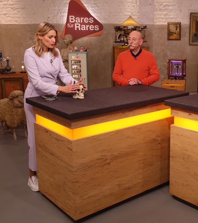 Expertin Dr. Bianca Berding (links) hat einen Bierkrug in Form eines Totenkopfes vor sich auf dem Tisch stehen und betrachtet ihn. Horst Lichter (Mitte) und der Verkäufer (rechts) schauen ihr zu.