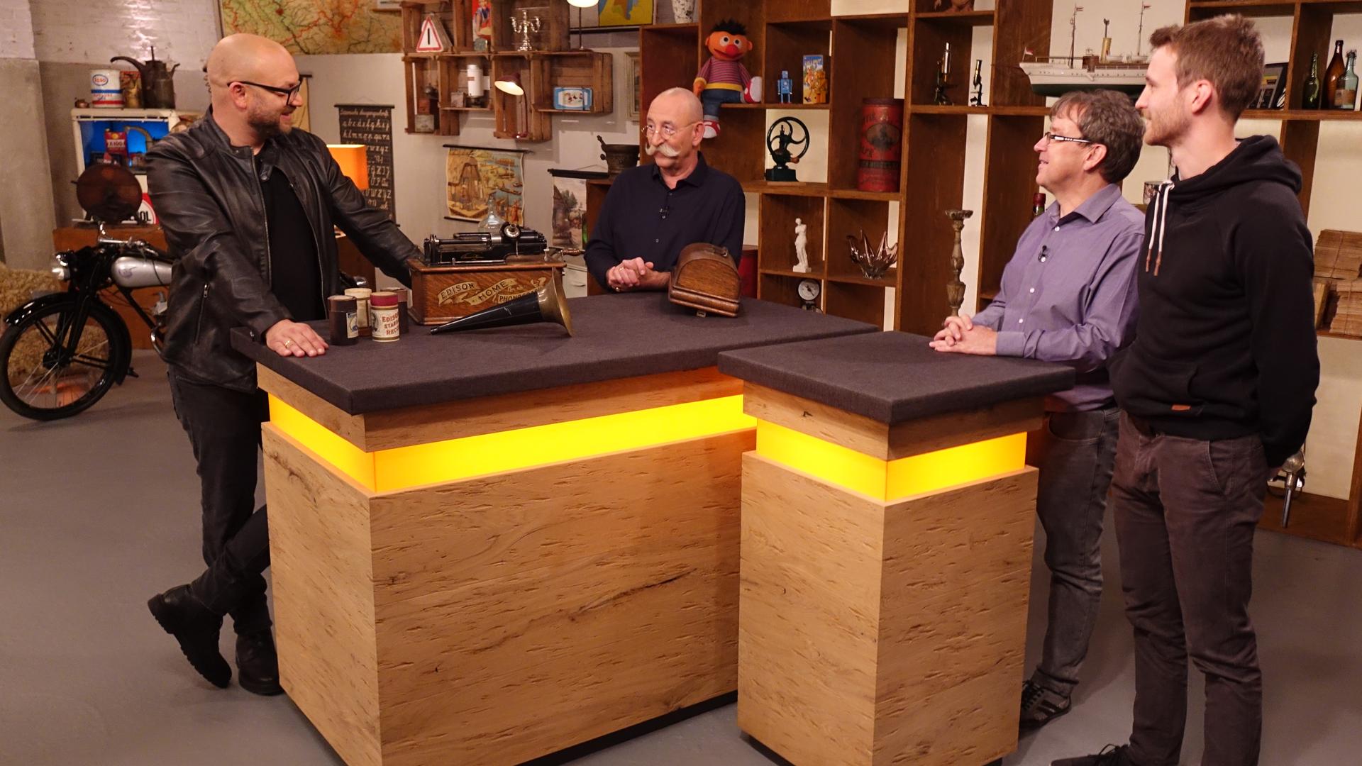 Experte Sven Deutschmanek (links) hat einen Phonographen vor sich auf dem Tisch stehen. Er spricht mit Horst Lichter (Mitte) und den beiden Verkäufern (rechts).