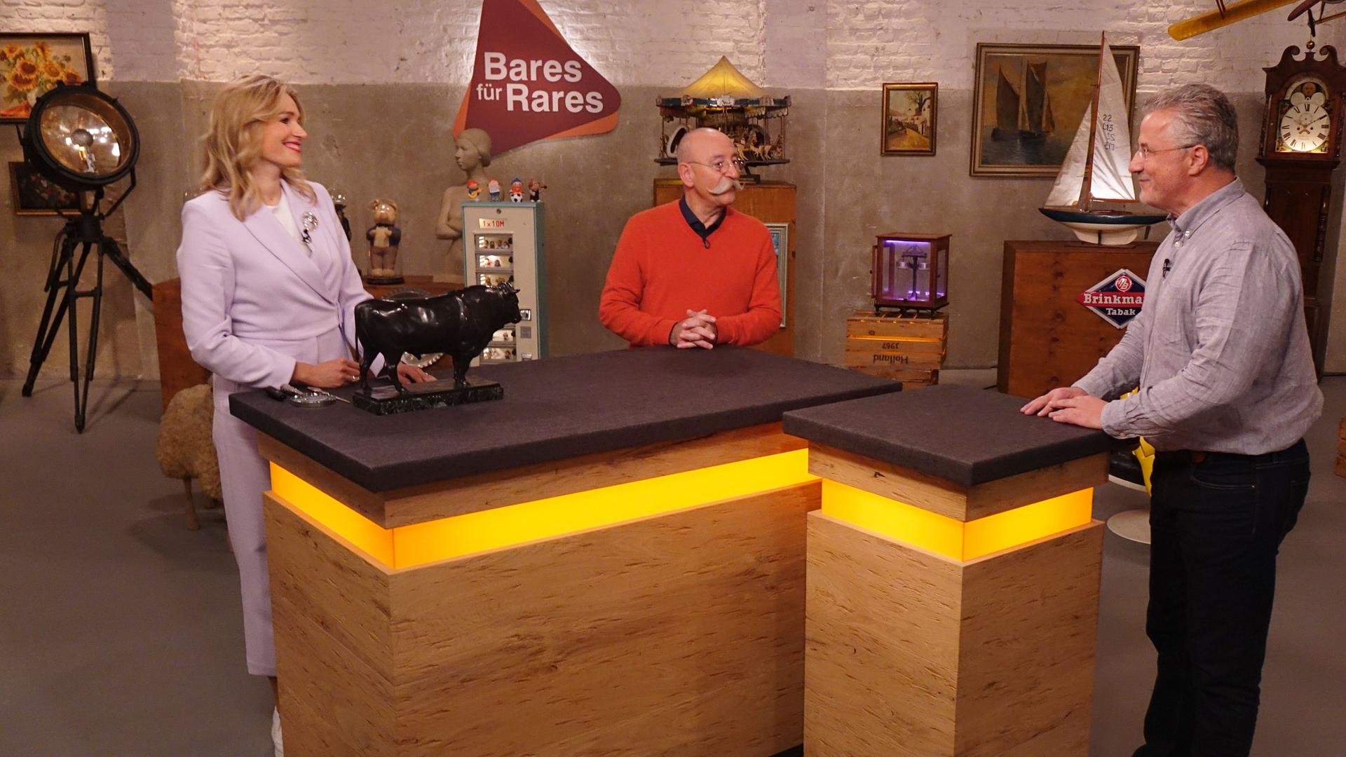 Expertin Dr. Bianca Berding (links) hat einen Bronze-Stier vor sich auf dem Tisch stehen. Sie und Horst Lichter (Mitte) schauen den Verkäufer (rechts) an.