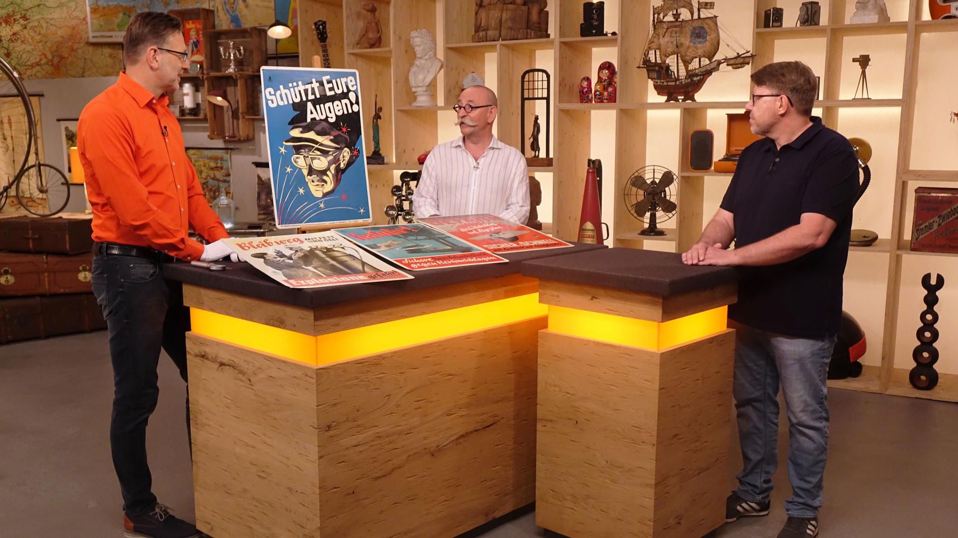 Experte Detlev Kümmel (links) steht neben dem Expertentisch, auf dem 4 Blechschilder platziert sind. Er spricht mit Horst Lichter (Mitte), der Verkäufer (rechts) schaut ihnen zu.