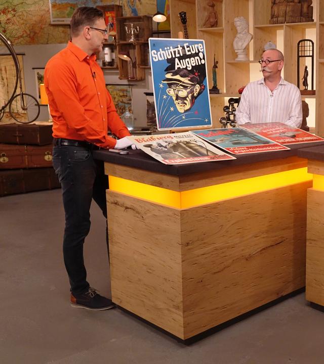 Experte Detlev Kümmel (links) steht neben dem Expertentisch, auf dem 4 Blechschilder platziert sind. Er spricht mit Horst Lichter (Mitte), der Verkäufer (rechts) schaut ihnen zu.