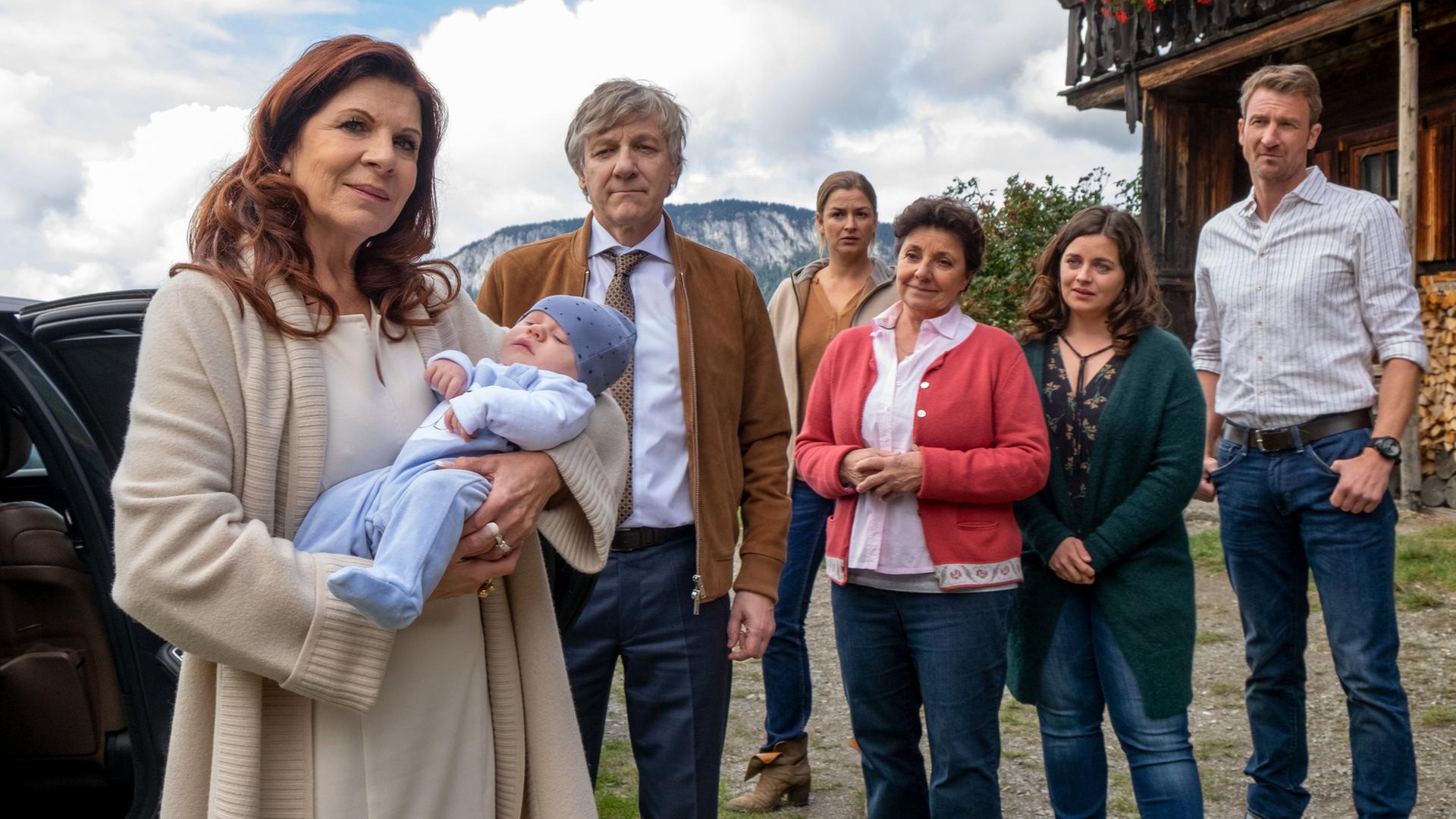 "Der Bergdoktor: Extreme": Vor dem Gruberhof: Thea Hochstetter (Claudia Wenzel) hält ein Baby auf dem Arm und lächelt. Im Hintergrund stehen Dr. Herold (Lutz Blochberger), Anne (Ines Lutz), Lisbeth (Monika Baumgartner), Lilli (Ronja Forcher) und Hans (Heiko Ruprecht) und blicken zu den beiden.